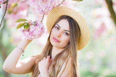 Pembe çiçekli güzel bir kadının bahar portresi. Uzun saçlı, hasır şapkalı ve kiraz çiçeklerinin yanında açık renk elbisesi olan güzel bir manken..