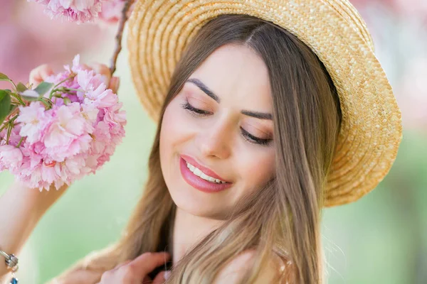 Spring portrait of a young beautiful woman in pink blossoms. Young beautiful model with long hair, in a wicker hat and a light dress near the cherry blossoms. — Fotografia de Stock