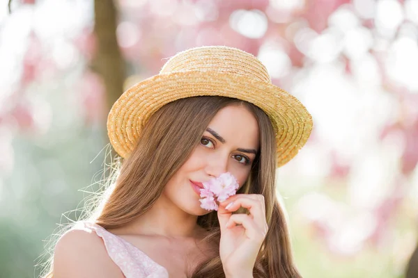 Pembe çiçekli güzel bir kadının bahar portresi. Uzun saçlı, hasır şapkalı ve kiraz çiçeklerinin yanında açık renk elbisesi olan güzel bir manken..