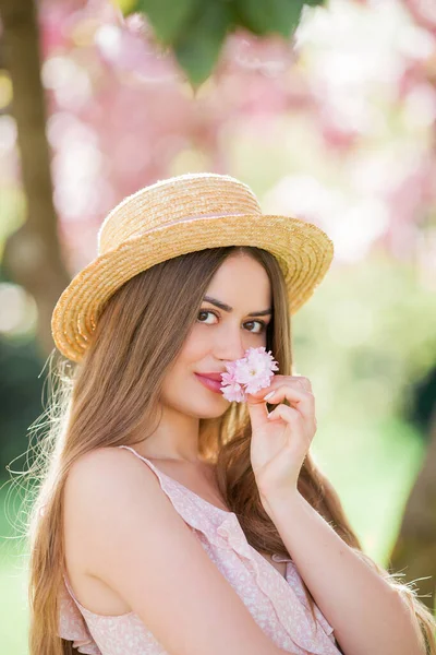 Vårporträtt av en ung vacker kvinna i rosa blommor. Ung vacker modell med långt hår, i en korgkeps och en lätt klänning nära körsbärsblommor. — Stockfoto
