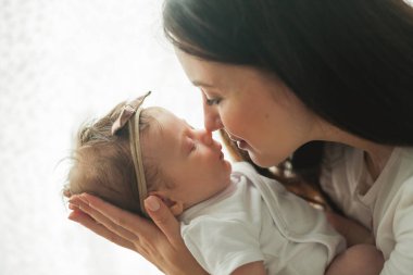 Genç, güzel, siyah saçlı anne yeni doğmuş kızını sıcak bir evde kollarında tutuyor. Aile fotoğrafı. Annelik. Genç bir kadın 2 aylık bebeğini öpüyor..