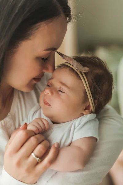 Young beautiful dark-haired mother holds her newborn daughter in her arms in a cozy home. Family portrait. Motherhood. Young woman kisses her 2 month old baby. — Fotografia de Stock