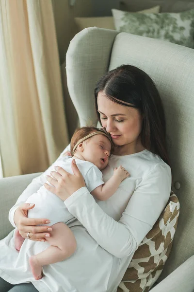 Mladá krásná tmavovlasá matka drží svou novorozenou dceru v náručí v útulném domově. Rodinný portrét. Mateřství. Mladá žena líbá ji 2 měsíce staré dítě. — Stock fotografie