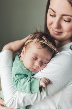 Genç, güzel, siyah saçlı anne yeni doğmuş kızını sıcak bir evde kollarında tutuyor. Aile fotoğrafı. Annelik. Genç bir kadın 2 aylık bebeğini öpüyor..