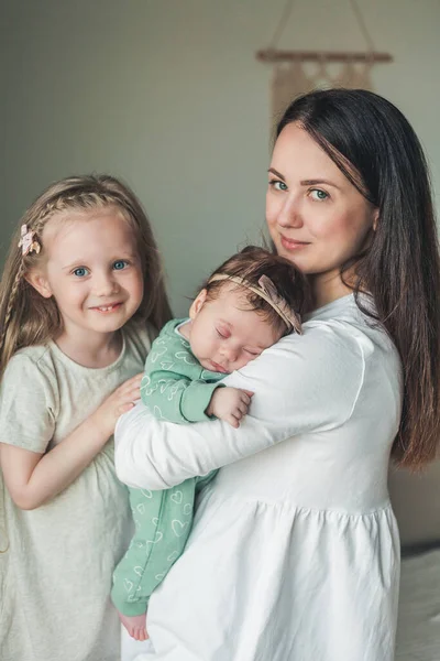 Young beautiful dark-haired mother in light clothes with her little daughters in a cozy house. Little sisters. Girls. Family. Happy motherhood. — Stock fotografie