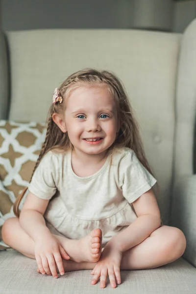 Una piccola ragazza dagli occhi azzurri carina 4 anni con i capelli lunghi è felice seduta in una poltrona in una casa accogliente. — Foto Stock
