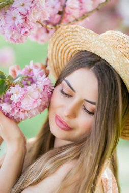 Pembe çiçekli güzel bir kadının bahar portresi. Uzun saçlı, hasır şapkalı ve kiraz çiçeklerinin yanında açık renk elbisesi olan güzel bir manken..