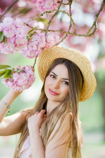 一个穿着粉色花朵的年轻漂亮女子的春照 年轻美丽的模特 头戴柳条帽 身穿轻薄的衣服 在樱花旁绽放 — 图库照片