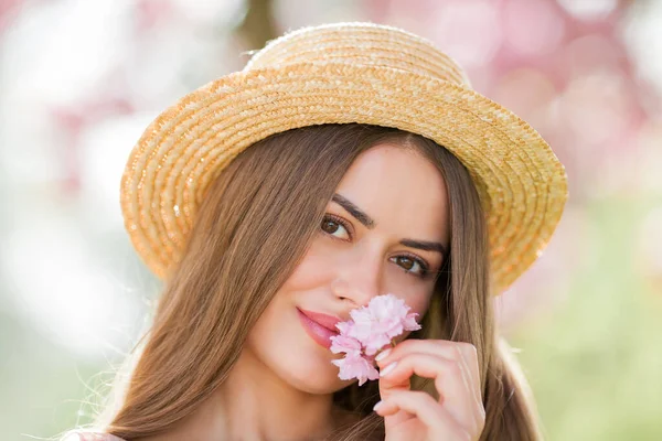 Spring portrait of a young beautiful woman in pink blossoms. Young beautiful model with long hair, in a wicker hat and a light dress near the cherry blossoms. — Stok fotoğraf