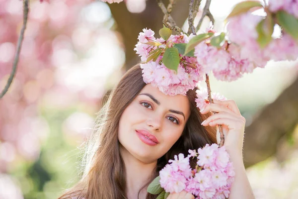 Pembe çiçekli güzel bir kadının bahar portresi. Uzun saçlı, hasır şapkalı ve kiraz çiçeklerinin yanında açık renk elbisesi olan güzel bir manken..