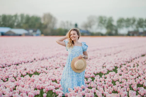 Beautiful young curly happy woman in pink tulip field in holland at sunset. Stylish model with a light romantic dress in a blooming field. Spring.