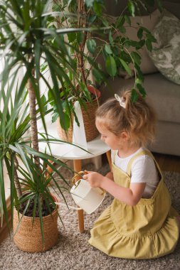 4 yaşındaki tatlı mavi gözlü kıvırcık saçlı kız sıcacık bir evdeki sulama kutusundan suyla çiçekleri döküyor. Küçük çocuk ev işlerinde annesine yardım ediyor. Çevre dostu. Gezegen. Doğa. Ev.