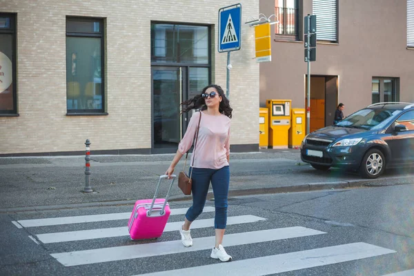 Beautiful young curly woman with a pink small suitcase is in a hurry to go on vacation. A model with long hair crosses the road in a European city.