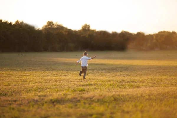 stock image Cute fair-haired boy of 5 years old in light simple clothes runs at sunset in a field in the sun. Happy child. Summer evening.