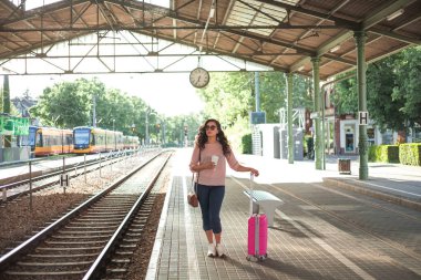 Pembe küçük bavullu 30-40 yaşlarında güzel bir kadın istasyonda trenini bekliyor. Günlük kıyafetleriyle telefonda konuşan, paket kahve içen kıvırcık bir manken. Seyahat.