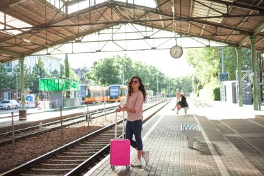 Pembe küçük bavullu 30-40 yaşlarında güzel bir kadın istasyonda trenini bekliyor. Günlük kıyafetleriyle telefonda konuşan, paket kahve içen kıvırcık bir manken. Seyahat.
