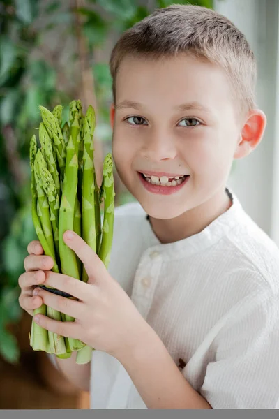 Cute happy fair-haired boy 7 years old holds a bunch of green juicy fresh asparagus in his hands. Healthy food. Eco product.