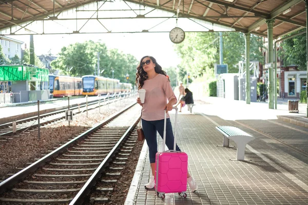 Pembe küçük bavullu 30-40 yaşlarında güzel bir kadın istasyonda trenini bekliyor. Günlük kıyafetleriyle telefonda konuşan, paket kahve içen kıvırcık bir manken. Seyahat.