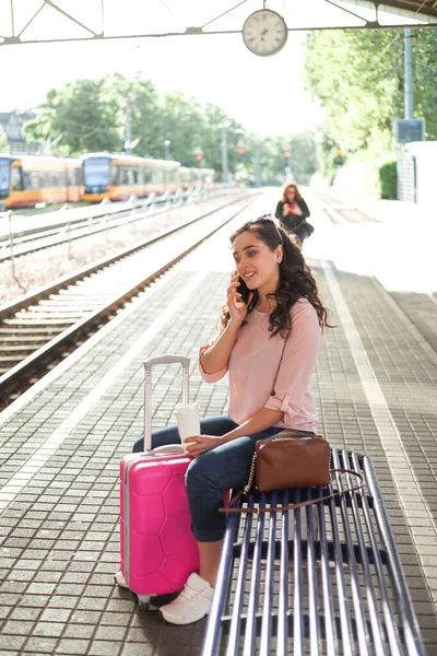 Young beautiful woman of 30-40 years old with a pink small suitcase is waiting for her train at the station. Curly model with casual clothes talking on the phone, drinking coffee to go. Traveling.