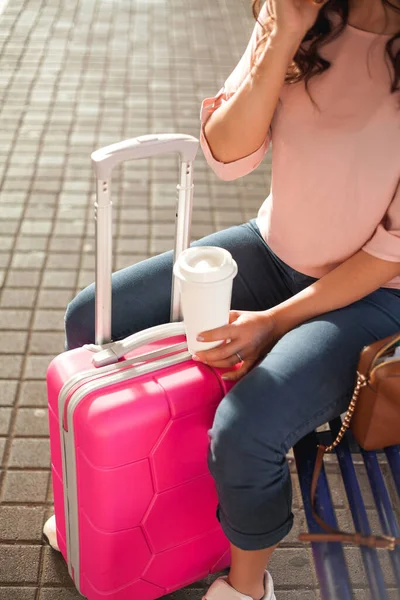 Young beautiful woman of 30-40 years old with a pink small suitcase is waiting for her train at the station. Curly model with casual clothes talking on the phone, drinking coffee to go. Traveling.