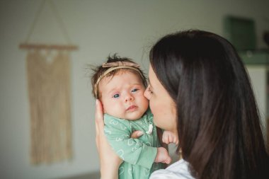 Genç, güzel, siyah saçlı anne yeni doğmuş kızını sıcak bir evde kollarında tutuyor. Aile fotoğrafı. Annelik. Genç bir kadın 2 aylık bebeğini öpüyor. Ev.