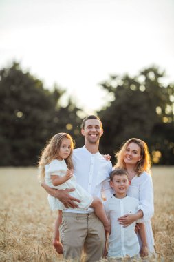 Beautiful young family at sunset in a wheat field. Happy parents with two children, a boy and a girl, in light clothes. Summer. Love.
