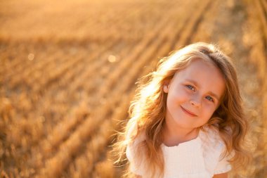 Adorable little curly girl 4 years old in a white dress in the sun at sunset in a mowed field of wheat. Happy child outside. Walk. Warm summer.
