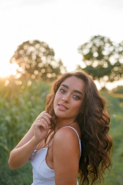 Delightful young woman in a white sundress in a field of green corn in the sunset light. Portrait of a beautiful model with dark long curly hair. Summer. Harvest. Eco. Bio.