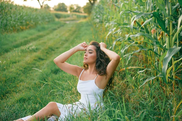 Delightful young woman in a white sundress in a field of green corn in the sunset light. Portrait of a beautiful model with dark long curly hair. Summer. Harvest. Eco. Bio.