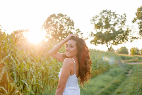 Delightful young woman in a white sundress in a field of green corn in the sunset light. Portrait of a beautiful model with dark long curly hair. Summer. Harvest. Eco. Bio.