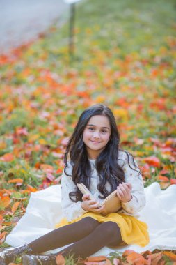 A pretty dark-haired 10-year-old girl in a knitted sweater reads a book sitting in a clearing in an autumn park. Picnic in nature. Happy child is learning. Fall.