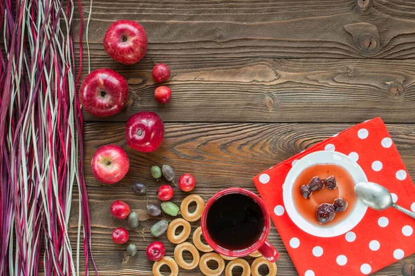 Kırmızı çay, olgun kırmızı elmalar, aktinidia üzümleri, simit, beyaz çay tabağında çilek reçeli, kaşık, ahşap masadaki beyaz polka noktalarında kırmızı peçete ve dekoratif renkli kuru pipet. en üstteki görünüm