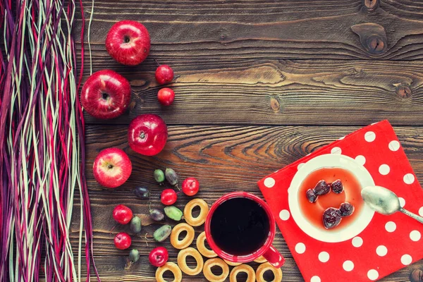 Kırmızı çay, olgun kırmızı elmalar, aktinidia üzümleri, simit, beyaz çay tabağında çilek reçeli, kaşık, ahşap masadaki beyaz polka noktalarında kırmızı peçete ve dekoratif renkli kuru pipet. en üstteki görünüm