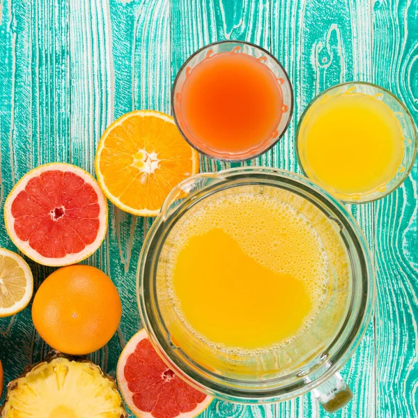 fresh juice from citrus fruits - lemon, grapefruit, orange, pine apple  in blender bowl and glasses on  turquoise colored wooden background, top view. copy space, free space for your text.