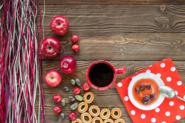 Kırmızı çay, olgun kırmızı elmalar, aktinidia üzümleri, simit, beyaz çay tabağında çilek reçeli, kaşık, ahşap masadaki beyaz polka noktalarında kırmızı peçete ve dekoratif renkli kuru pipet. en üstteki görünüm