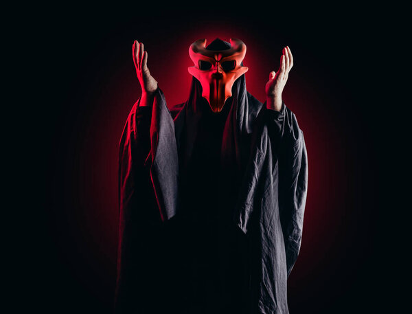 Scary horror occult sectarian priest in black hood and metal mask on black background with red glow.