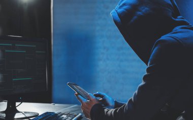 Karanlık odada bilgisayar ve cep telefonuyla yazı yazan bir bilgisayar korsanı şifreleri hacklemek için cep telefonuyla kötü amaçlı yazılım kullanıyor kişisel veri bankadan para çalıyor. siber