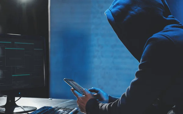 Накладные хакеры в капюшоне, работающие за компьютером и мобильным телефоном, печатающие текст в темной комнате, анонимный хакер использует вредоносные программы с мобильным телефоном, чтобы взломать пароль, личные данные крадут деньги из банка. кибер Лицензионные Стоковые Фото