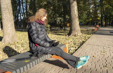 Sonbahar parkında bankta oturan ve akıllı telefonuna bakan genç bir kadın. Parkta sonbahar havası var..