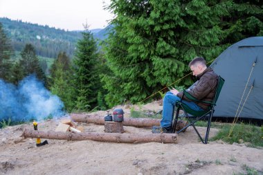 Genç adam ormanda kamp çadırının yanında dinleniyor.