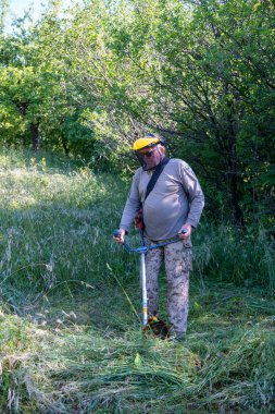 Koruyucu maskeli olgun bir adam bahçede çimleri buduyor.