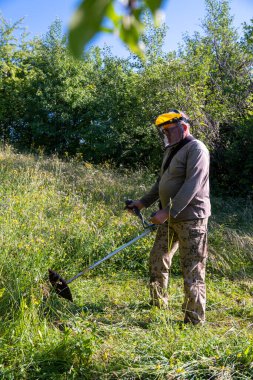 Koruyucu maskeli olgun bir adam bahçede çimleri buduyor.
