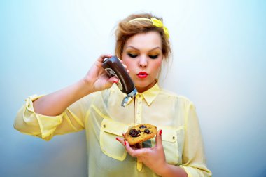 Young stylish woman with red hair and a yellow bow headband wearing red lipstick and a yellow chiffon blouse is pouring chocolate sauce over chocolate chip cookie. clipart