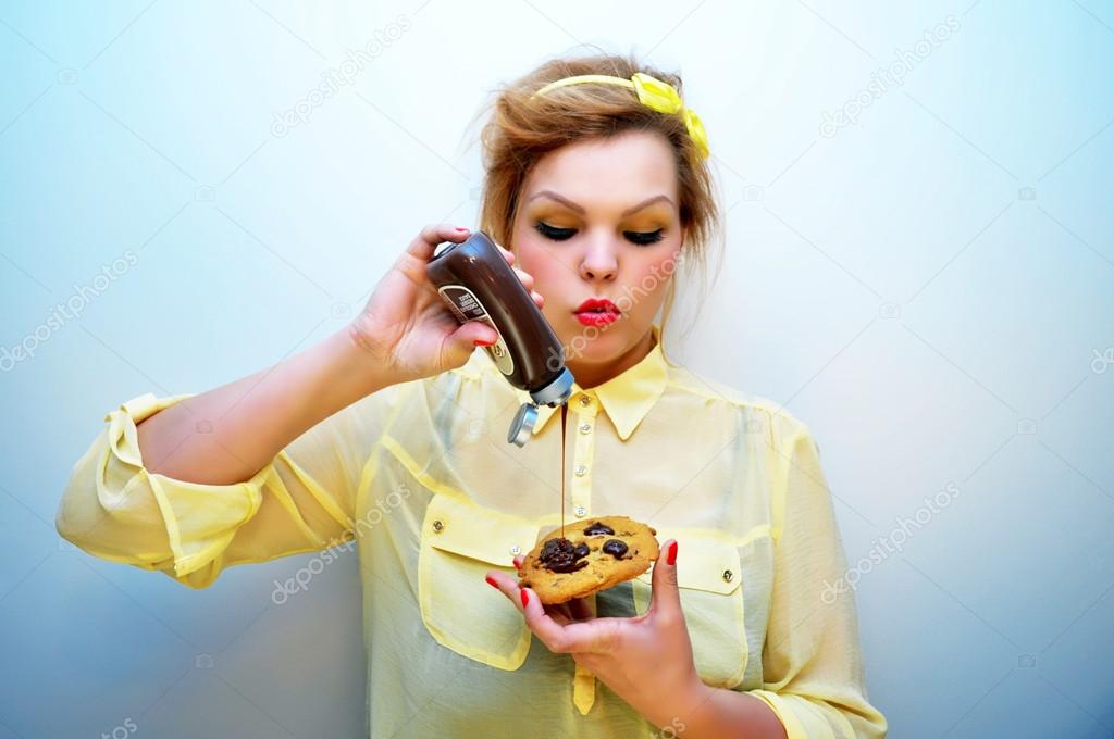 Young stylish woman with red hair and a yellow bow headband wearing red lipstick and a yellow chiffon blouse is pouring chocolate sauce over chocolate chip cookie.