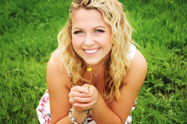 Smiling beautiful blonde young woman with curly hair wearing a flower print summer dress and holding a small yellow flower in her hands on a green meadow.