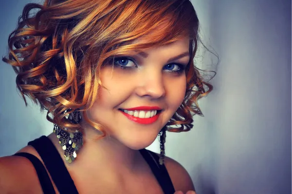 Smiling beautiful young woman with curly short red hair, red lipstick and evening make-up wearing big sparkling earrings. — Stock Photo, Image