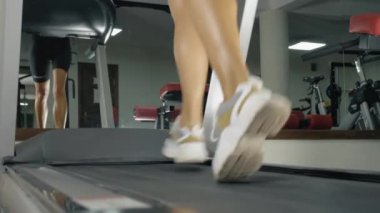 Bir kadın koşu bandında kardiyo ısınması yapıyor. Spor salonundaki genç bir kızın bacaklarının alçak açılı görüntüsü..
