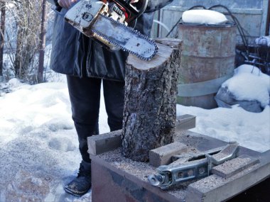 Kışın kırsal bir bahçede elektrikli testerenin ucuyla tahta bir kütüğü işlerken, bir ağaç gövdesinin bir parçası, bir kriko ile birlikte, benzin testereli bir adam tarafından işleme konuldu.