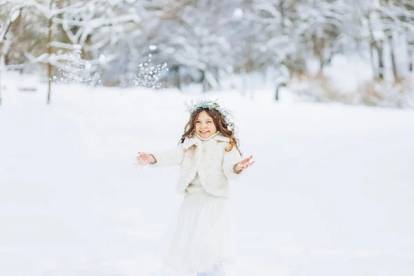 雪を投げ雪の中に立って 冬の公園で 花輪と雪で遊んで白いコートで幸せな小さな巻き女の子 晴れた日 クリスマス — ストック写真