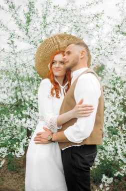 Beyaz çiçeklerin arka planında kucaklaşan sevgi dolu bir çift. Eski tarz, hasır şapkalı kız..